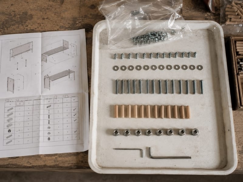 Flat-pack hardware bag emptied onto white tray: bolts sorted into rows, one row visibly short, printed kit list beside tray.